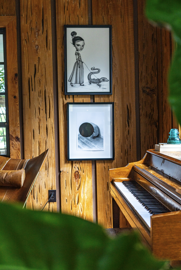 Wood-paneled wall with two framed art prints, a girl with a leash, and a snake; a brown leather chair; and an upright piano with a blue jar and books on top.