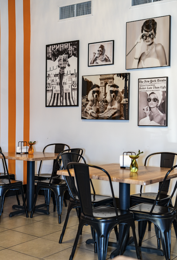 A cozy cafe interior with wooden tables, black metal chairs, small vases with flowers, and framed black-and-white photos on white walls, featuring fashion and daily life scenes.