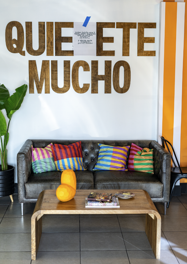 Decorative wall with large wooden letters spelling 'QUIETETE MUCHO' and a note attached with blue tape. A black tufted leather sofa with colorful striped pillows sits beneath the wall. A wooden coffee table in front of the sofa holds a yellow decorative object, magazines, and a plate. To the left, there is a tall green plant in a black pot. The floor is tiled, and there is a black chair or table on the right.