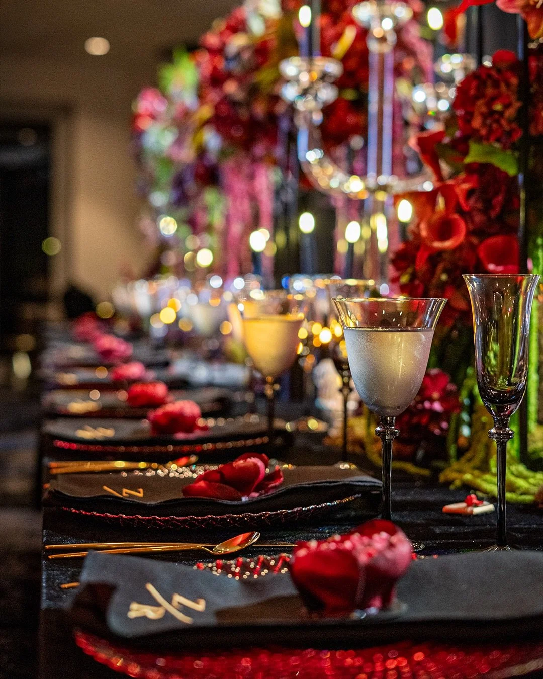 Elegant banquet table setting with black napkins, red rose petals, and gold cutlery, illuminated by soft lighting, with a bouquet of red flowers and hanging decorations in the background.