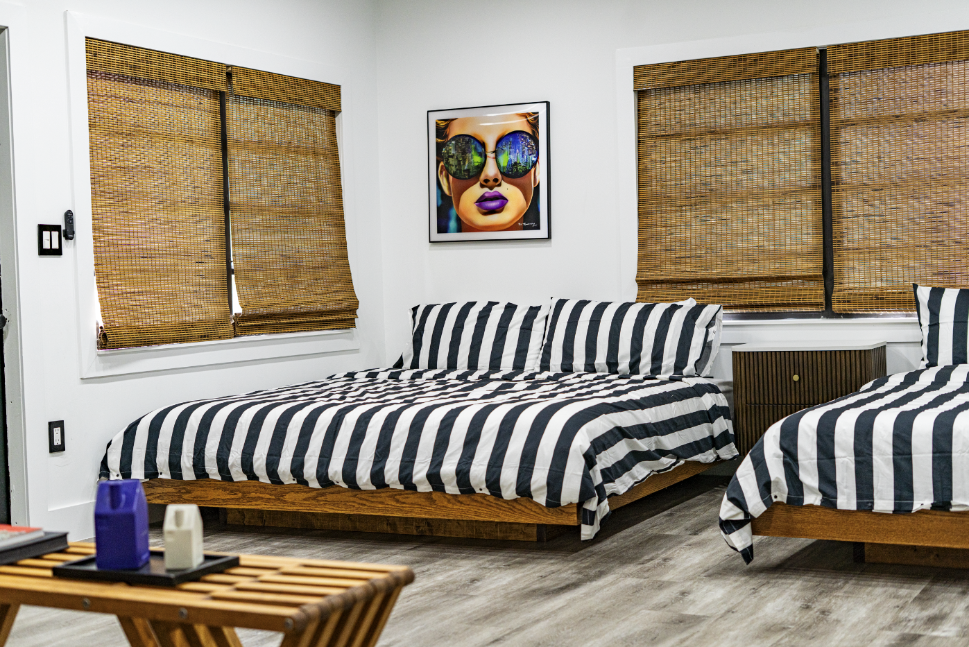 A bedroom with two beds covered in black and white striped bedding, wooden bed frames, and a wooden nightstand. There are two windows with bamboo blinds, a framed artwork of a woman wearing sunglasses, and a wooden table with candles in the foregroun