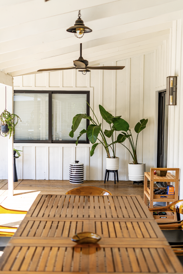 Indoor patio with wooden dining table, large potted plants, ceiling fan, hanging light, window, and white paneled walls.