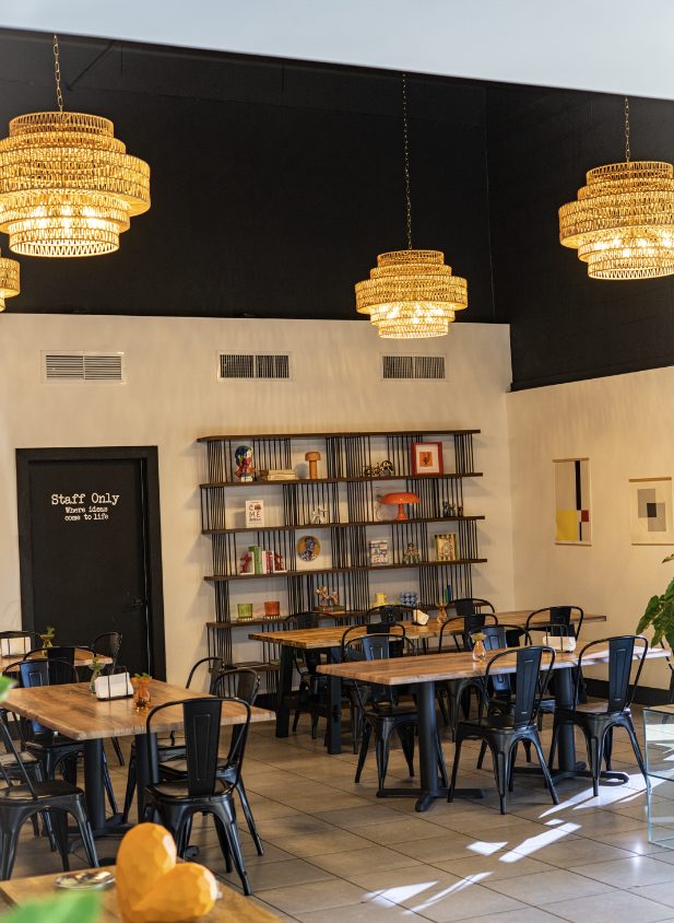 Interior of a restaurant or cafe with wooden tables, black chairs, a decorative shelf with books and ornaments, and three large, gold-colored chandeliers hanging from a black ceiling.