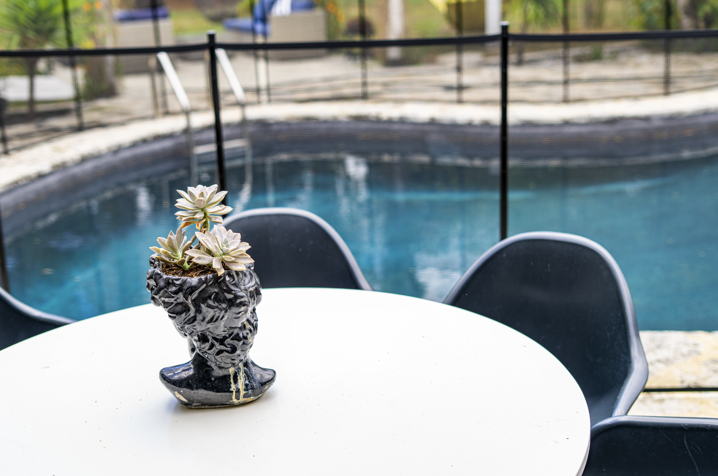 A black and white decorative bust-shaped planter with succulent plants, on a white outdoor table near a swimming pool, with poolside chairs in the background.