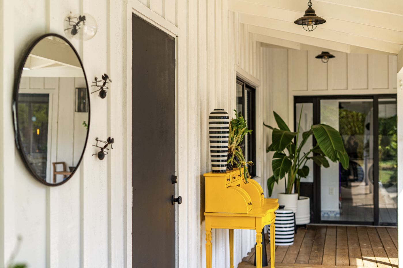 Interior of a bright porch with white paneled walls, a black door, a large potted plant, and a yellow piano with striped vase and potted plant on top, with a glass sliding door at the back.