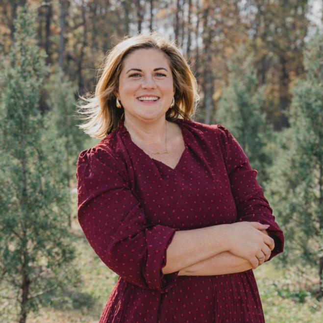 A woman with shoulder-length blonde hair smiling outdoors, wearing a burgundy dress with small polka dots, standing in front of trees and greenery.