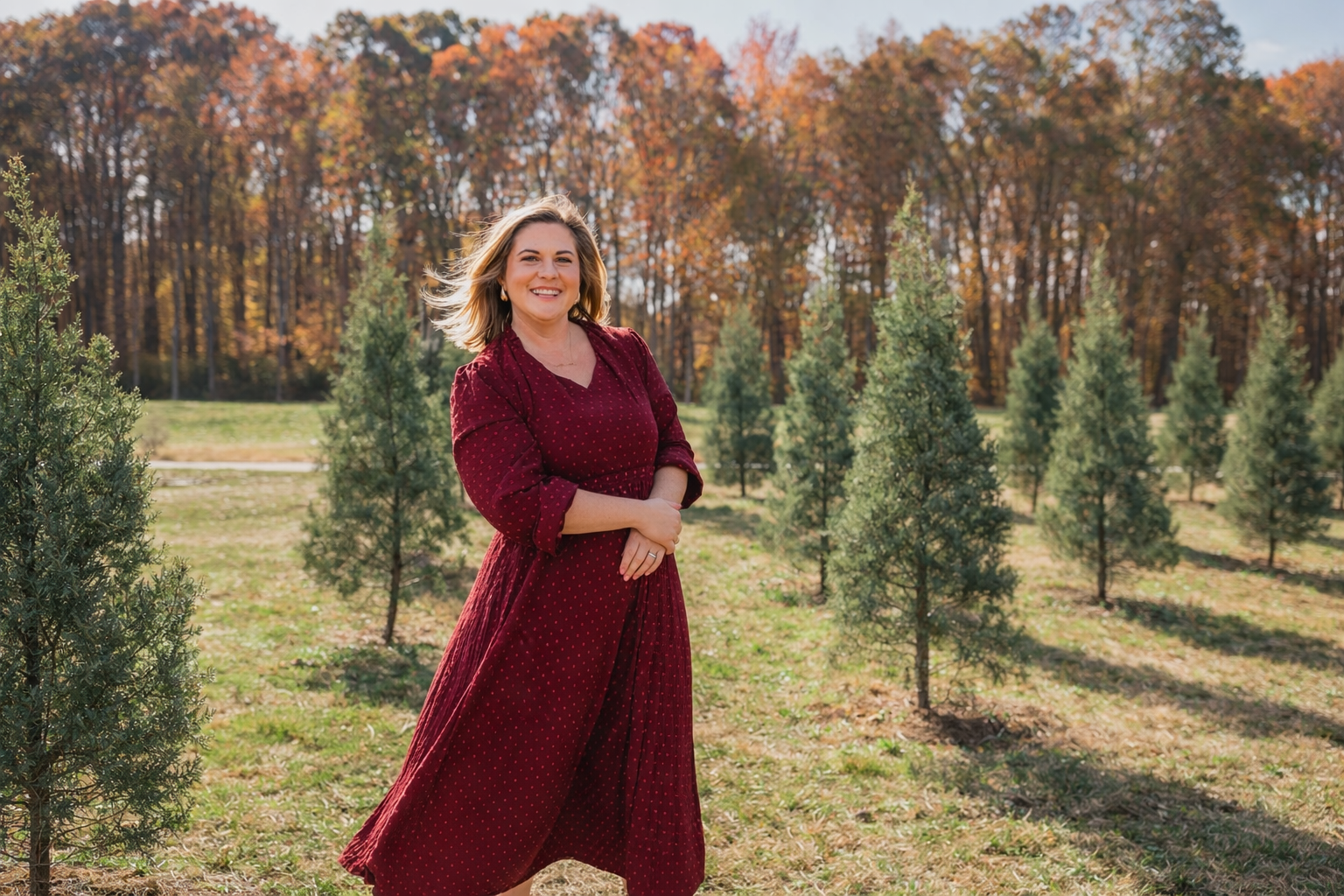 A woman wearing a red dress standing in a tree farm with small evergreen trees and fall foliage in the background.
