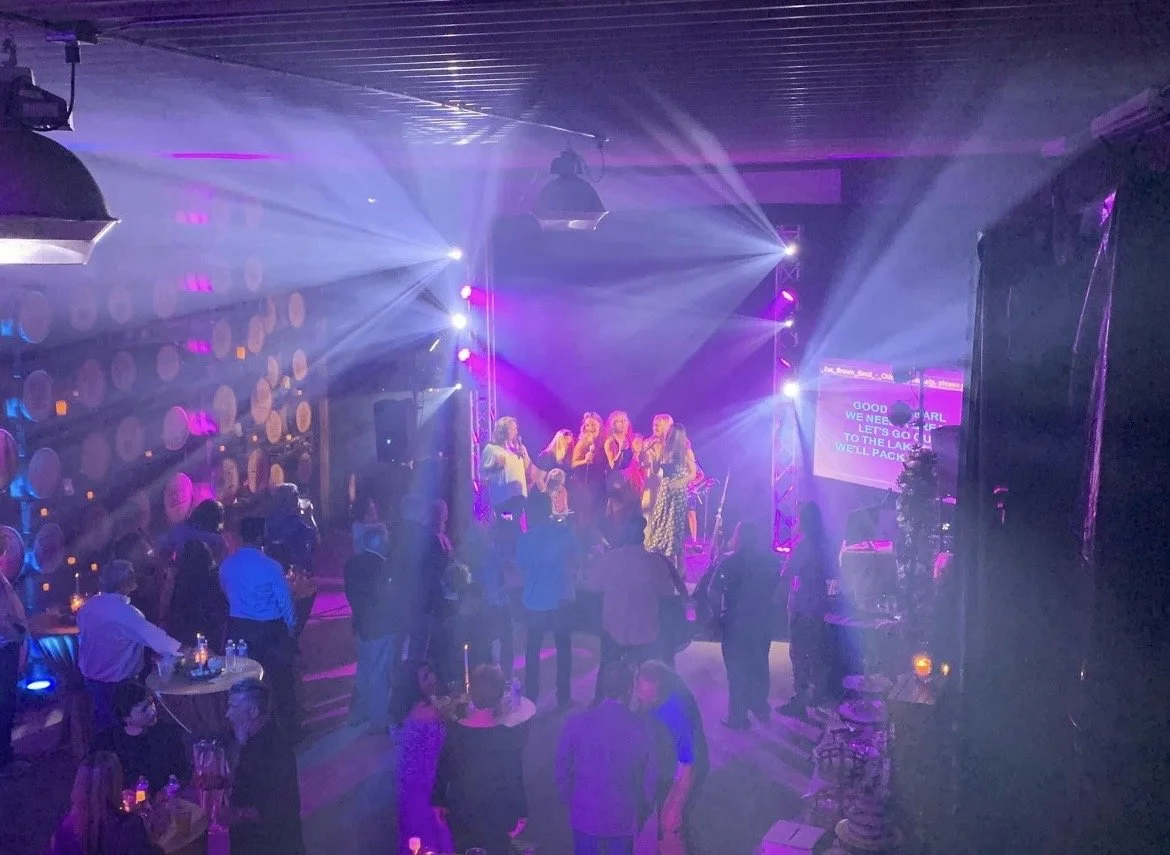 People dancing and socializing in a dimly lit nightclub with colorful lighting, a stage with performers or a karaoke screen, and a wall decorated with barrels.