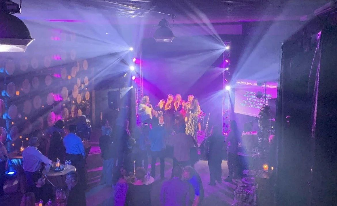 People dancing and socializing at a karaoke event in a dimly lit club with purple and blue lighting, stage with singers, and a screen displaying lyrics.