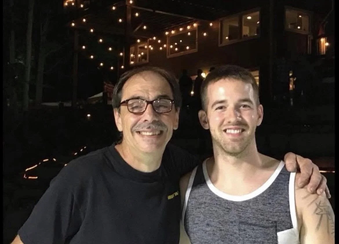 Two men smiling and posing in a nighttime outdoor setting with string lights and a house in the background.