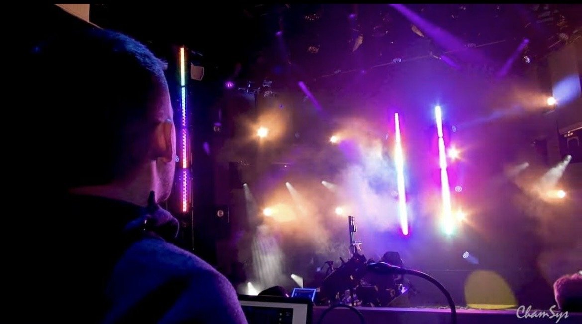 A person with glasses and a mask watching a colorful, lit stage at a concert or nightclub with vibrant purple, blue, and white lights and smoke effects.