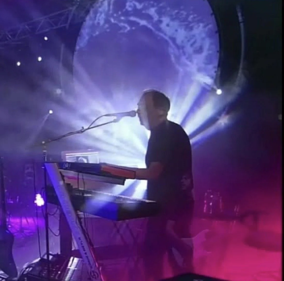 A man playing a keyboard and singing into a microphone on stage, with colorful stage lights and a large circular screen displaying a blue pattern behind him.