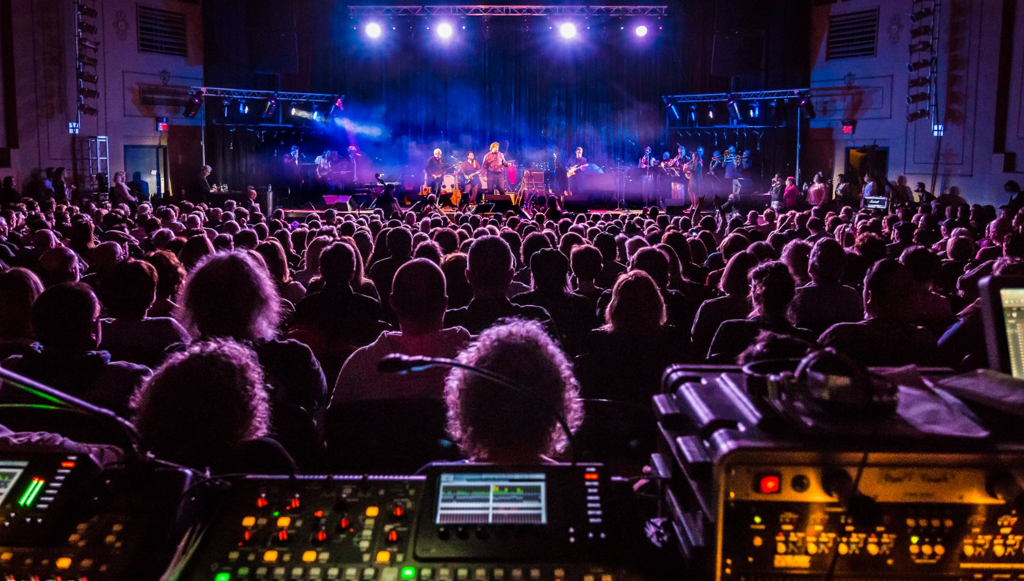 An indoor concert with an audience watching a band perform on stage, illuminated by purple and blue lights, with audio equipment in the foreground.