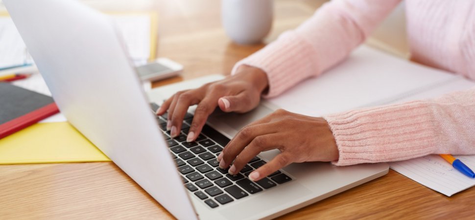 Person typing on a laptop keyboard at a wooden desk with notebooks, papers, and a pen.