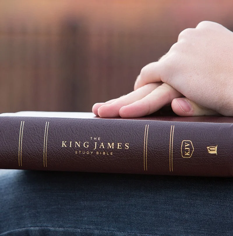 A person's hand resting on a closed, dark brown leather-bound King James Study Bible, with gold lettering and symbols on the cover.