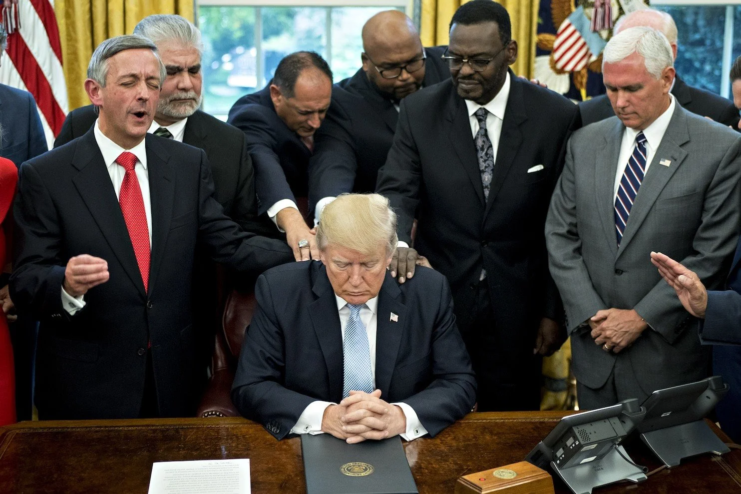 Donald Trump sitting at a desk with his hands clasped, surrounded by several men praying or paying respects in the Oval Office.