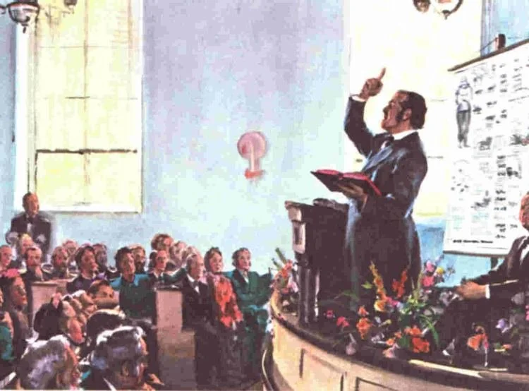 A man in a suit is standing at a podium speaking to an audience in a large room with blue walls and a window. He is holding a folder and gesturing with his right hand. There are several people seated in the audience, listening attentively. The room is decorated with flowers and a large displayed chart or document is behind the speaker.