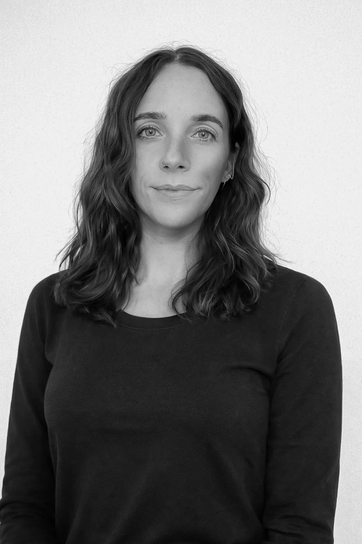 A woman with wavy shoulder-length hair, wearing a dark long-sleeve shirt, looking at the camera with a slight smile, standing against a plain light-colored background.