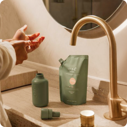 Hands using a bottle opener near a bottle on a bathroom countertop, with a soap dispenser and a refill pouch nearby.