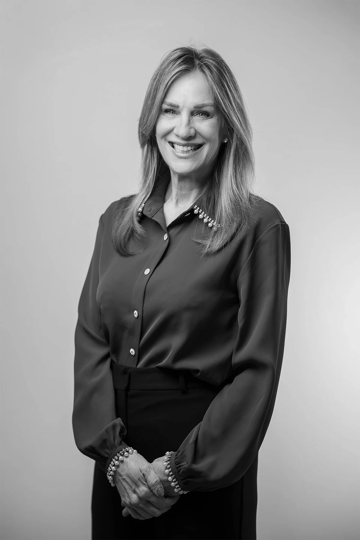 A black-and-white portrait of a smiling middle-aged woman with shoulder-length hair, wearing a dark blouse with decorative buttons and pearl earrings, standing against a plain background.