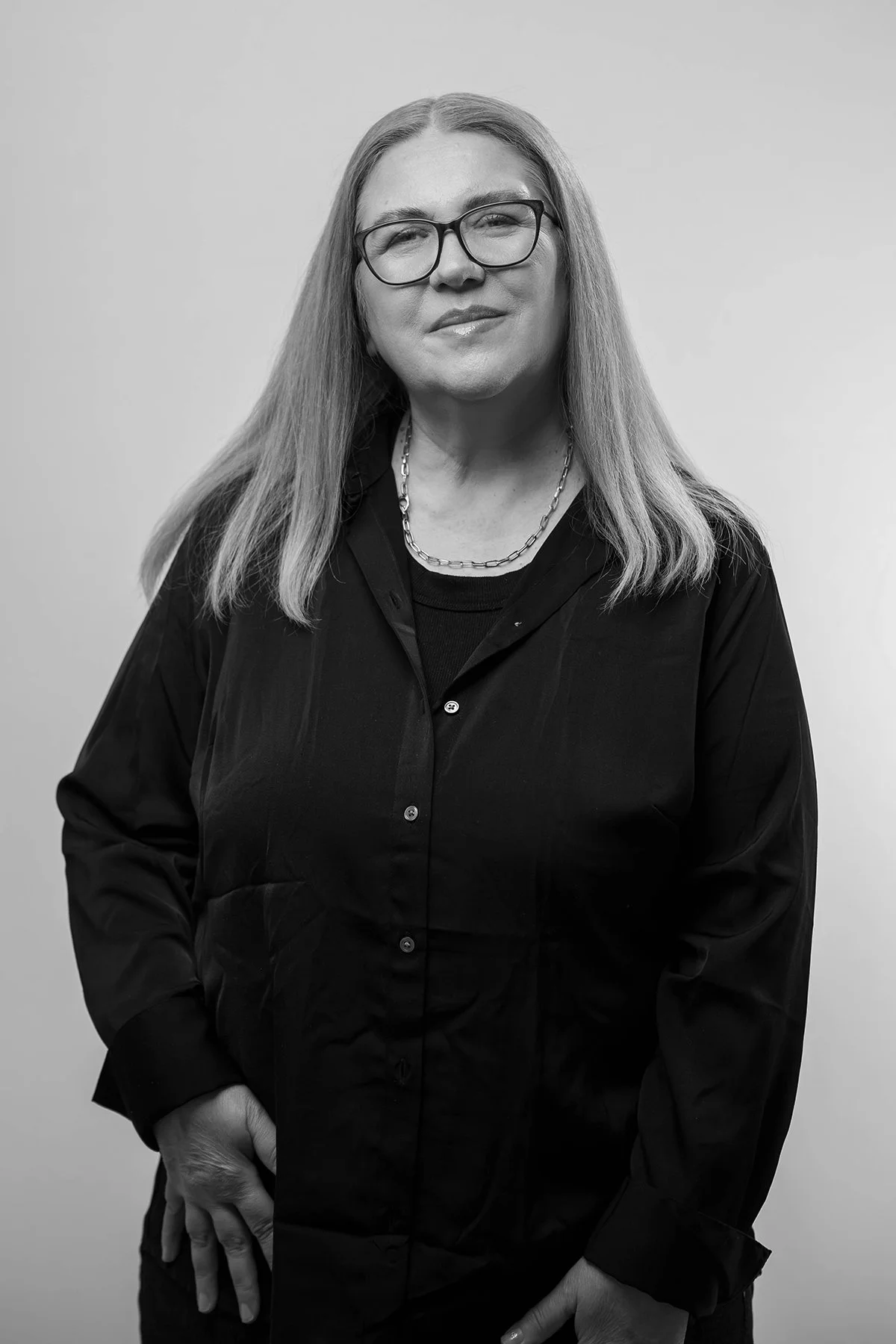 Black and white portrait of a woman with long hair, glasses, wearing a dark shirt and a chain necklace, standing against a plain background.