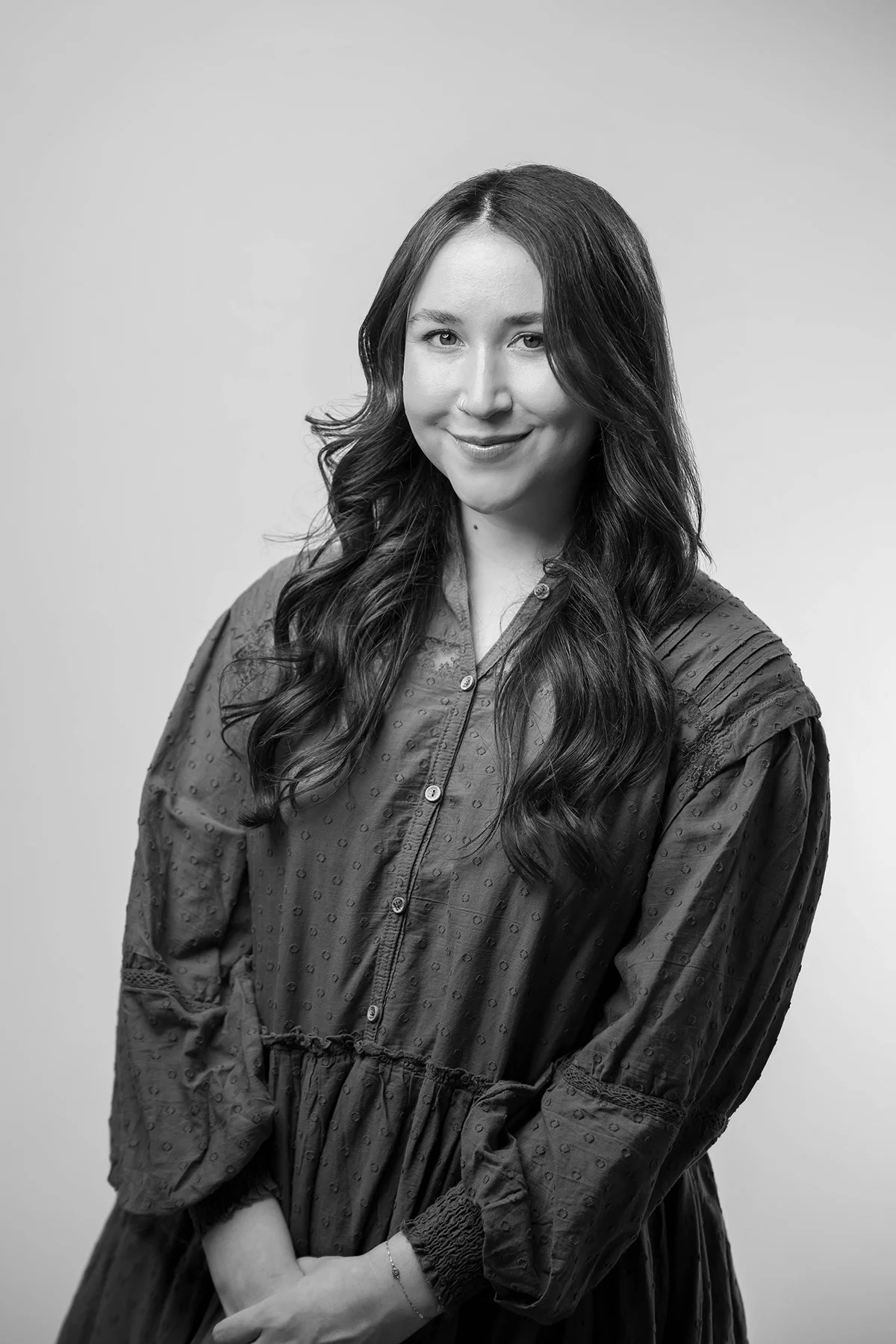 Black and white portrait of a young woman with long wavy hair, smiling slightly, wearing a textured button-up dress with loose sleeves.