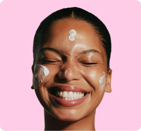 A woman with her eyes closed and a big smile on her face, with cream or lotion on her face, forehead, and cheeks, against a pink background.