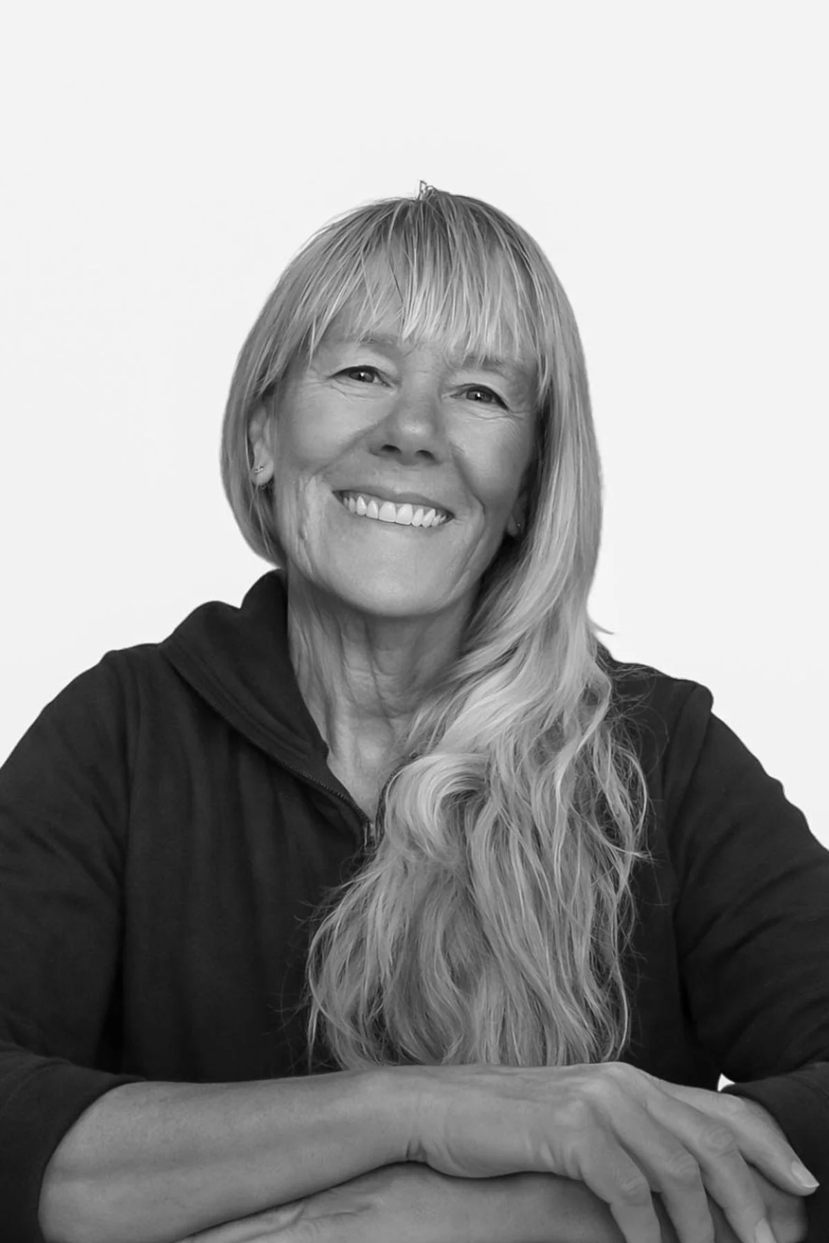 A black and white portrait of a smiling woman with long wavy hair, wearing a dark zip-up top, sitting with her arms crossed.