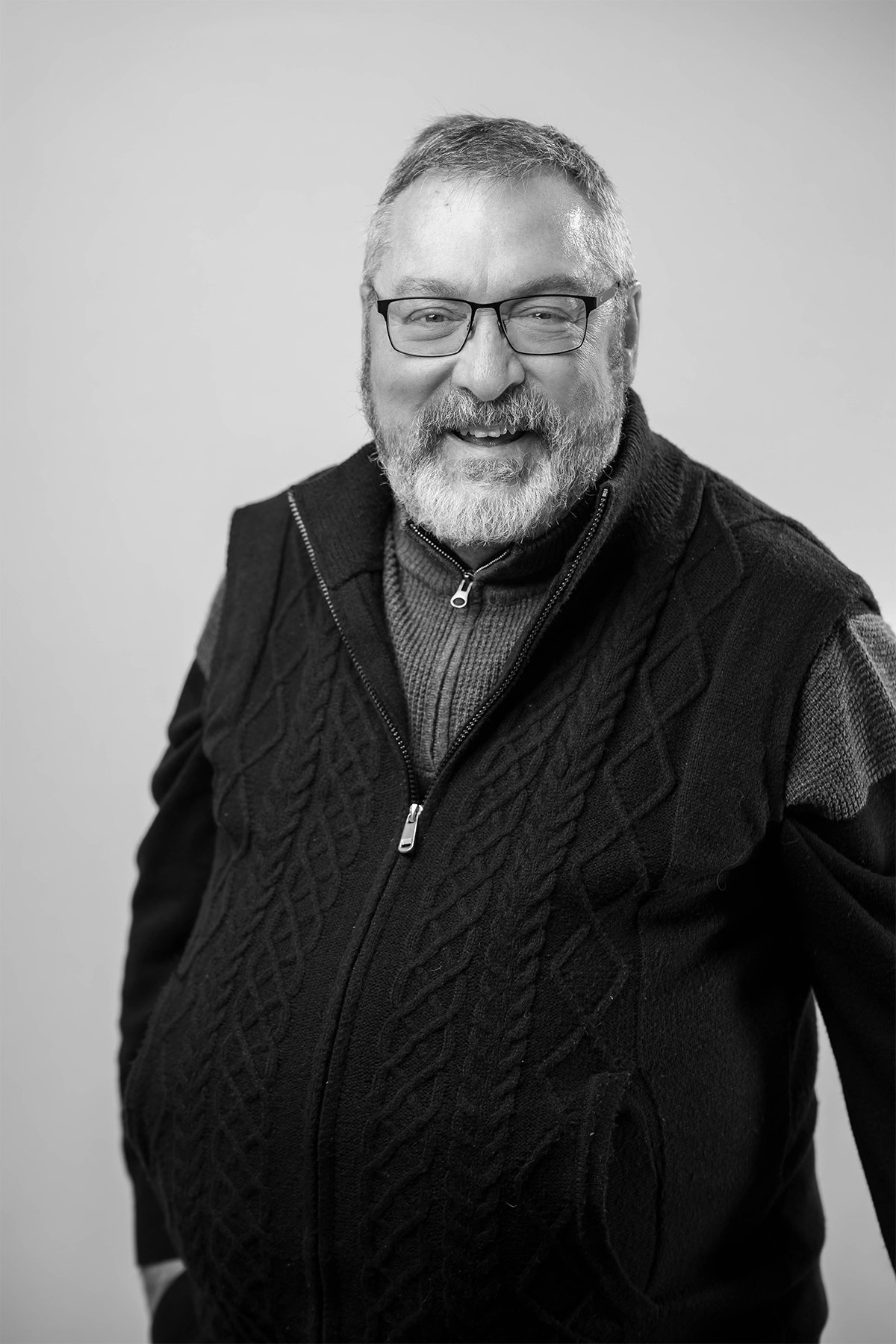 A smiling middle-aged man with glasses, a beard, and short hair, wearing a zip-up sweater with a pattern, standing against a plain background.
