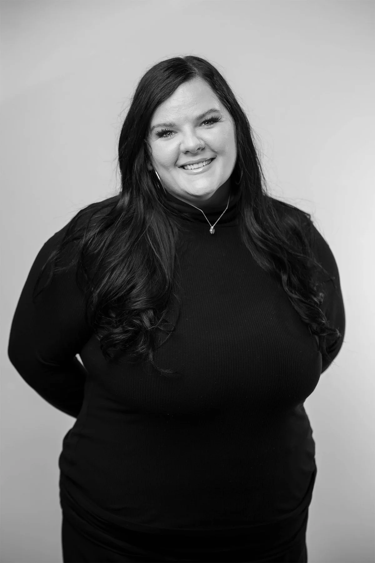 A smiling woman with long dark hair, wearing a black turtleneck and jewelry, standing against a plain background.