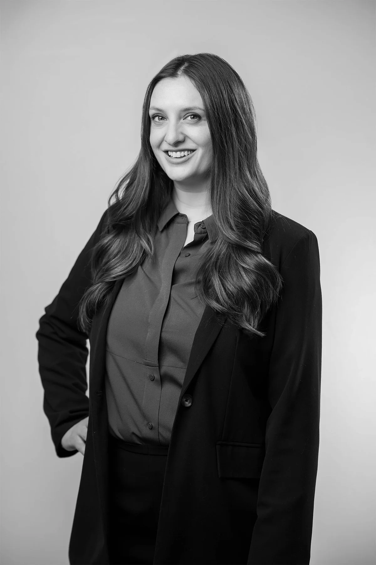 Black and white portrait of a woman with long wavy hair wearing a dark blazer over a button-up shirt, smiling with her hand on her hip against a plain background.