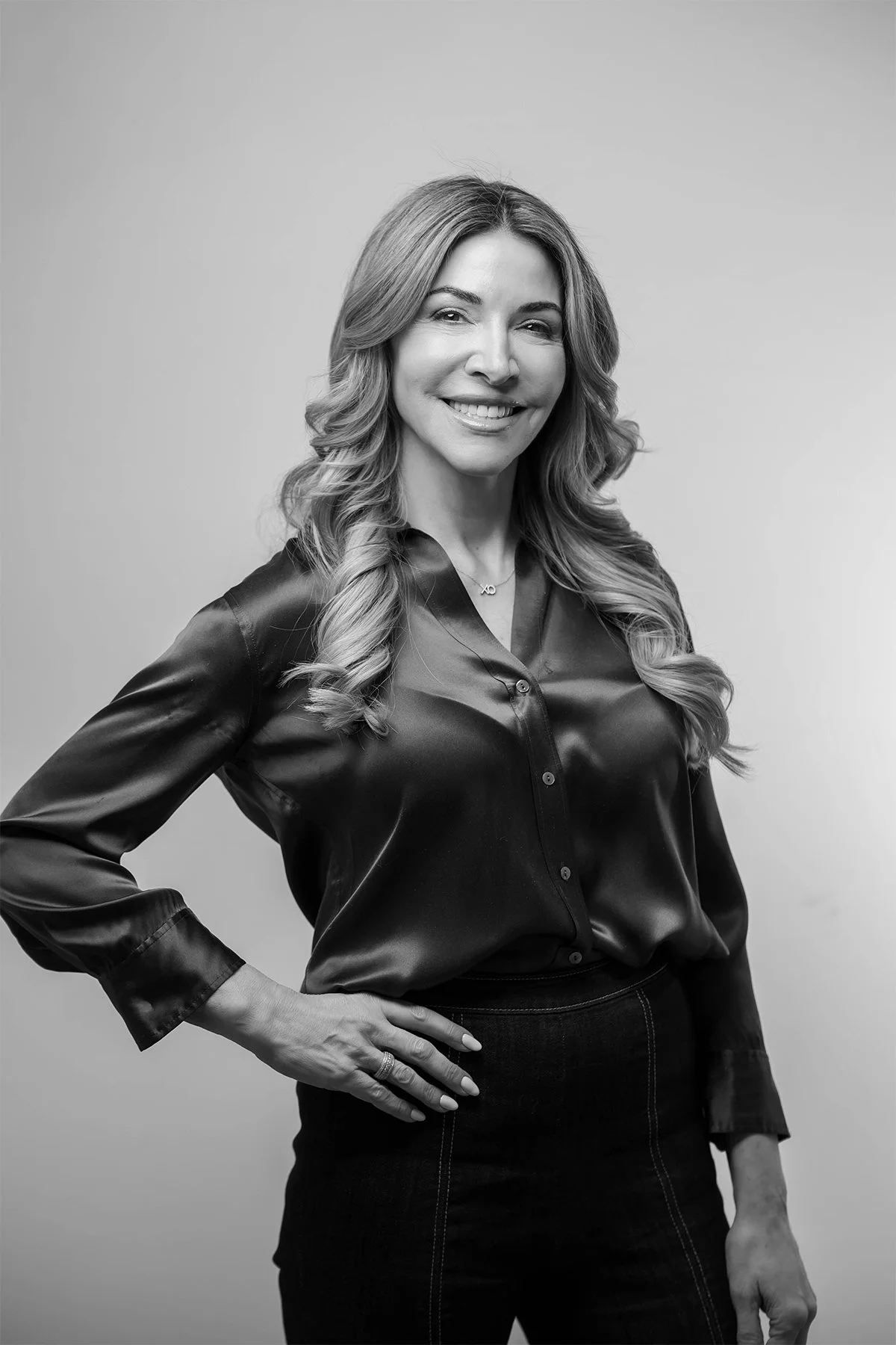 Black and white portrait of a woman with long wavy hair, wearing a shiny satin blouse and a dark skirt, smiling with one hand on her hip.
