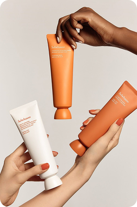 Two hands holding shampoo bottles, one white and one orange, against a neutral background.