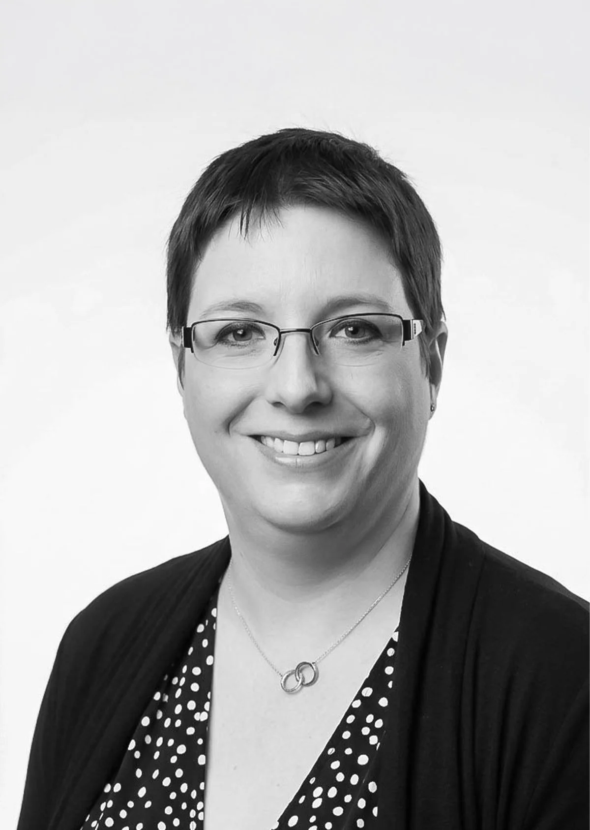 A black and white portrait of a woman with short hair, wearing glasses, a polka dot blouse, a black cardigan, and a necklace with two interlinked circles, smiling at the camera.