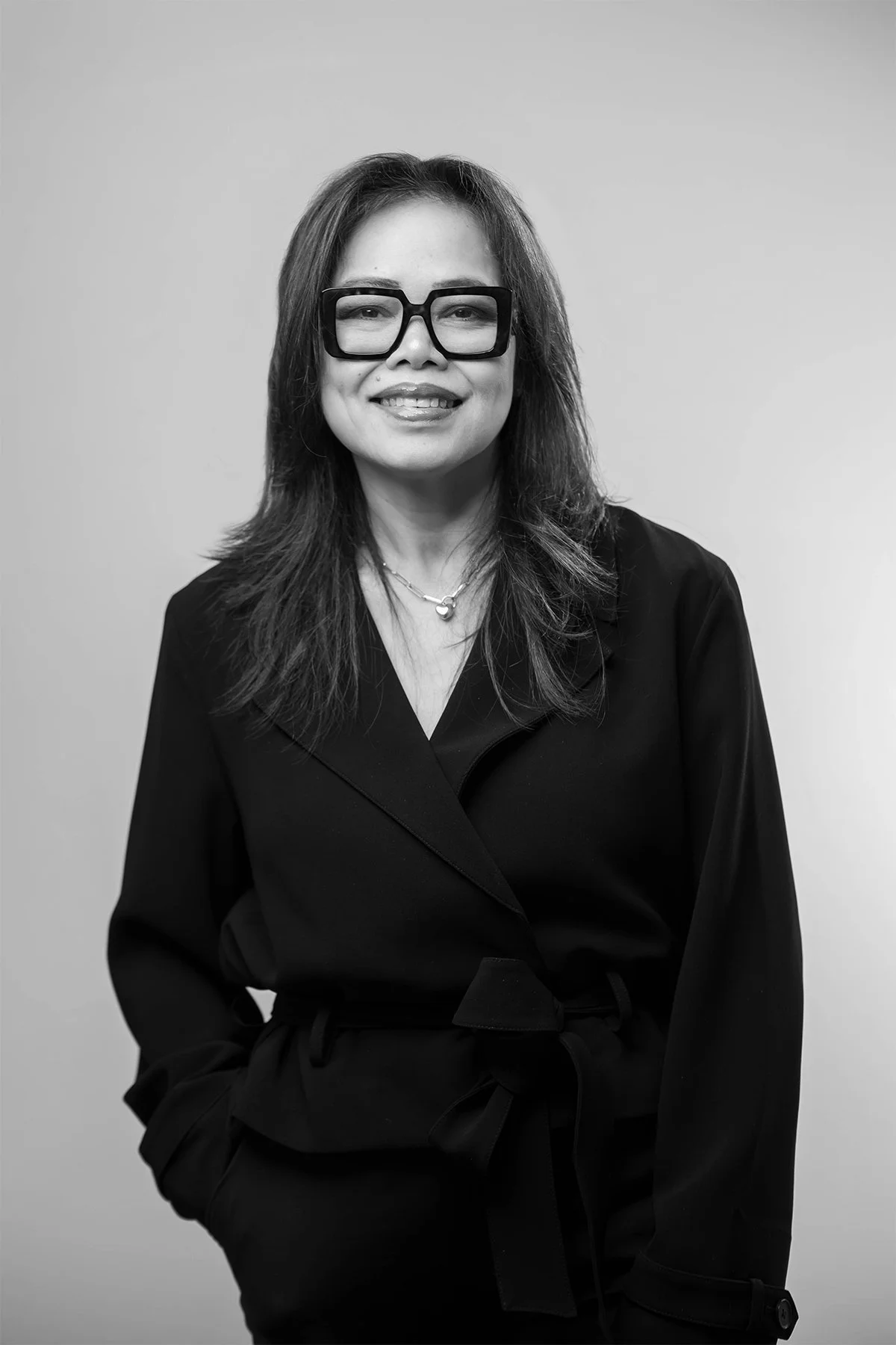 Black-and-white portrait of a woman with shoulder-length hair, glasses, smiling, wearing a dark long-sleeved wrap top with a belt, and layered necklaces, standing against a plain background.
