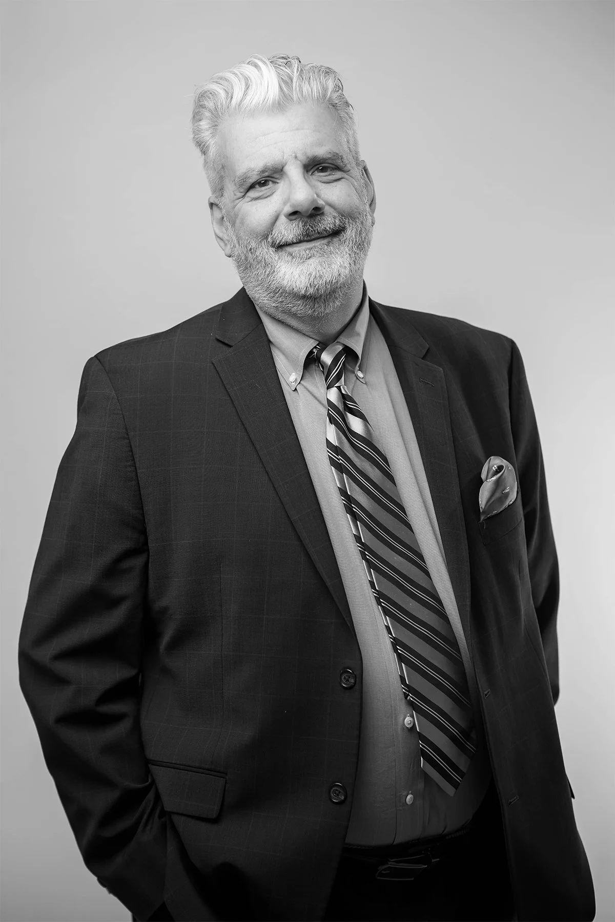 Black and white portrait of a smiling older man with gray hair and beard, wearing a dark suit, light shirt, striped tie, and pocket square, standing against a plain background.