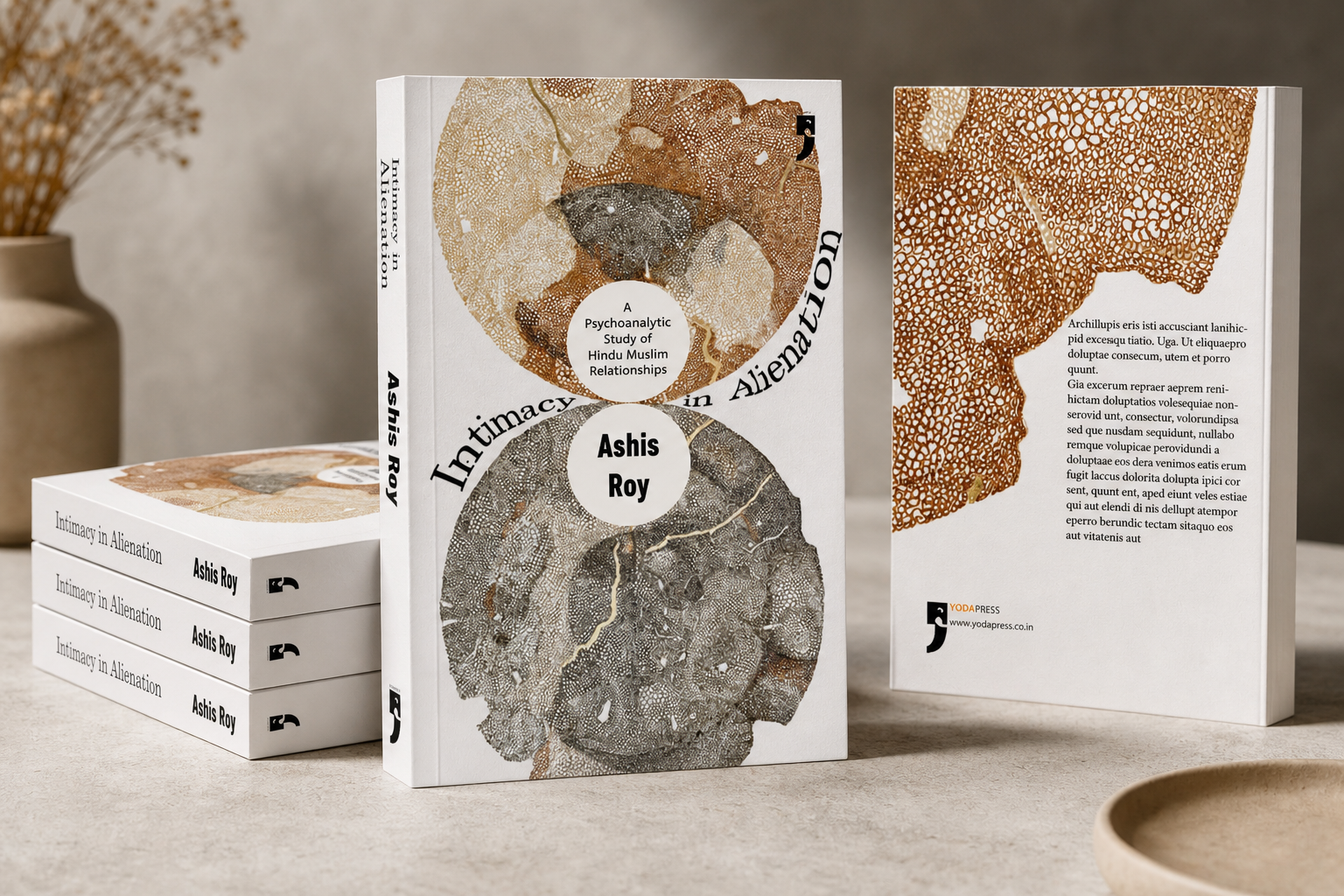 A stack of four white copies of the book titled "Intimacy in Alienation" by Ashis Roy on a table, with a single standing copy showing the front cover and back cover, featuring abstract textured circular designs in earthy tones, and a flat round wooden dish in the foreground and a vase with dried flowers in the background.