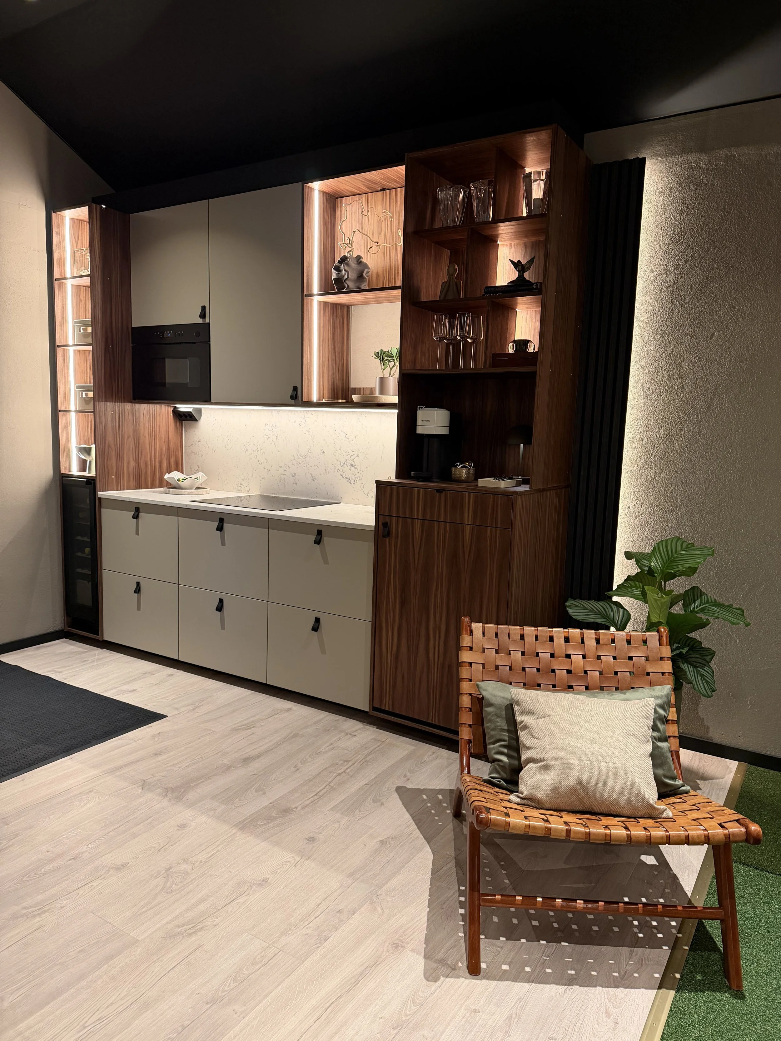 Ett modernt kök med vita skåp, trähylla och en sittplatsstol bredvid en växt.