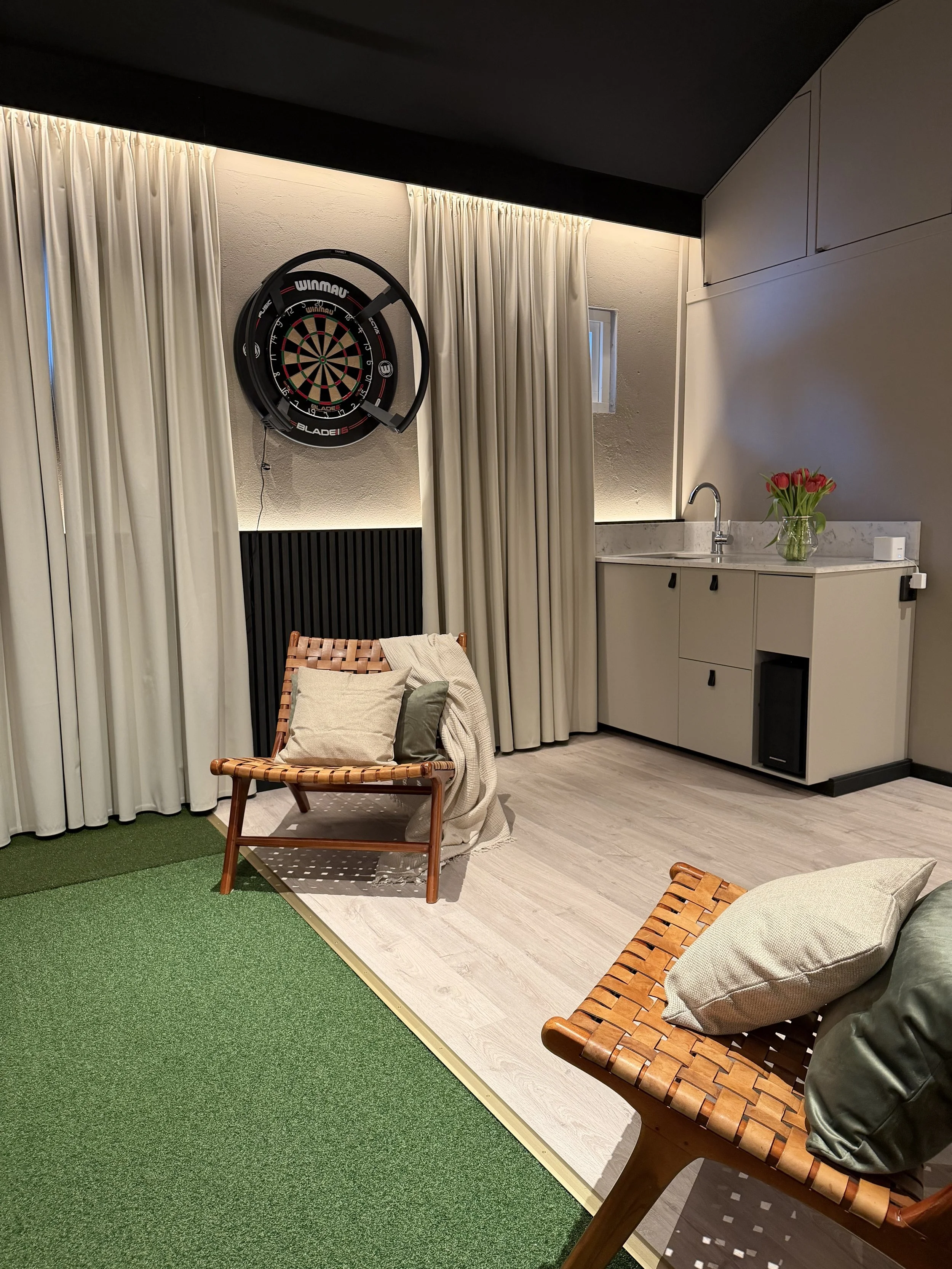 Ett rum med ett dartskåp, två vimplar, en skinnbänk med kuddar, en liten köksbänk med vask och blommor, ett grönt område och ett fönster.