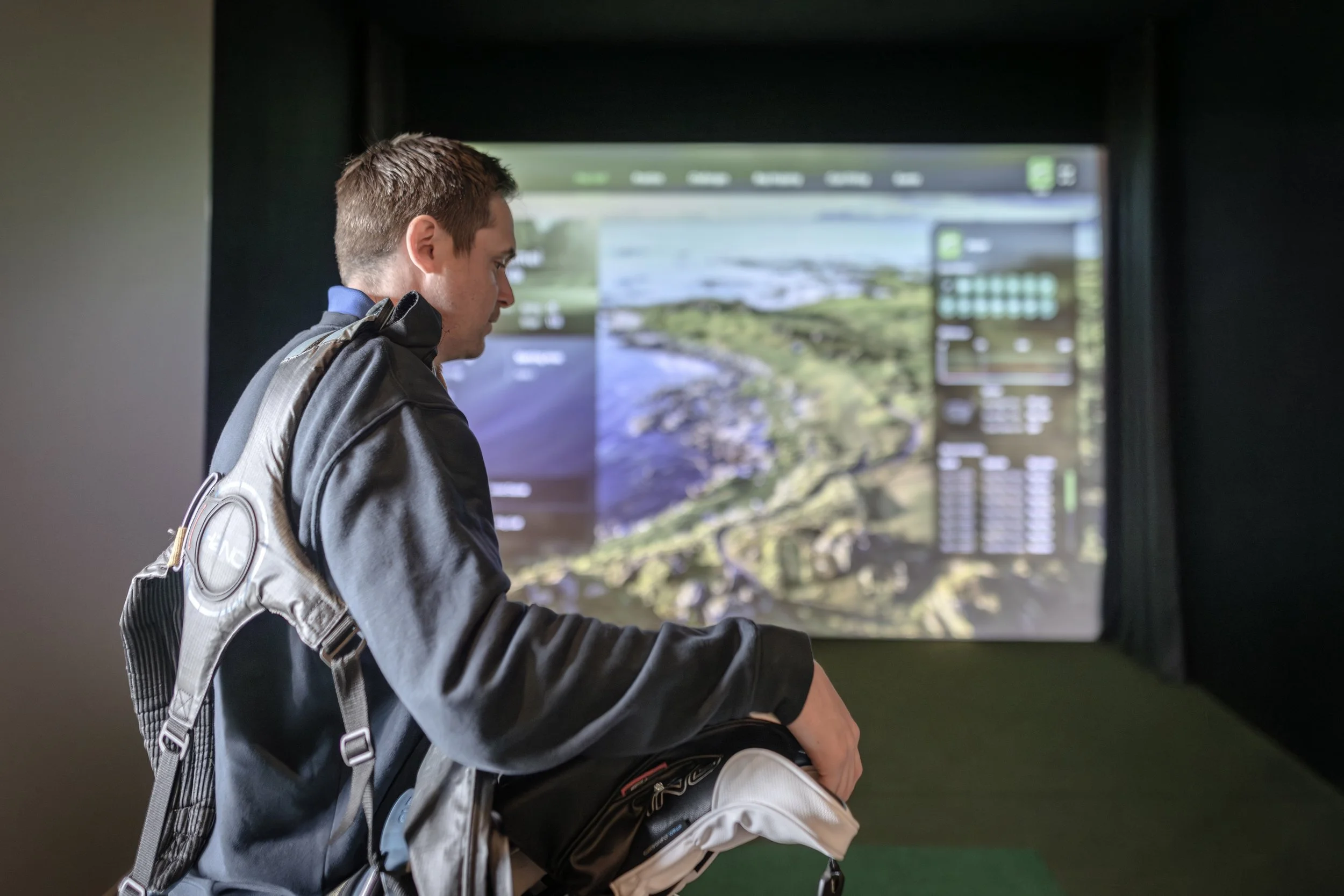 En ung man med ryggsäck tittar på en stor skärm som visar en landskapsbild med geodatainformation.