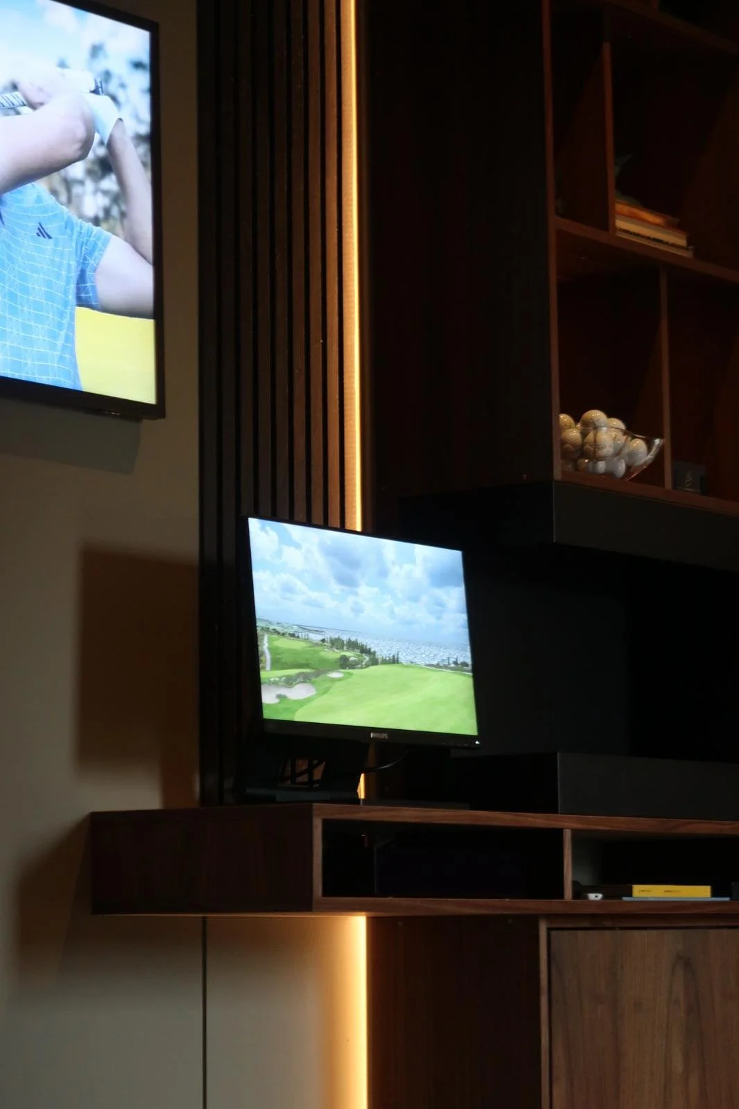 En inomhusmiljö med en vägg med träpaneler, två TV-skärmar, en av dem visar ett landskapsbilder av golfbana och den andra en person som spelar golf.