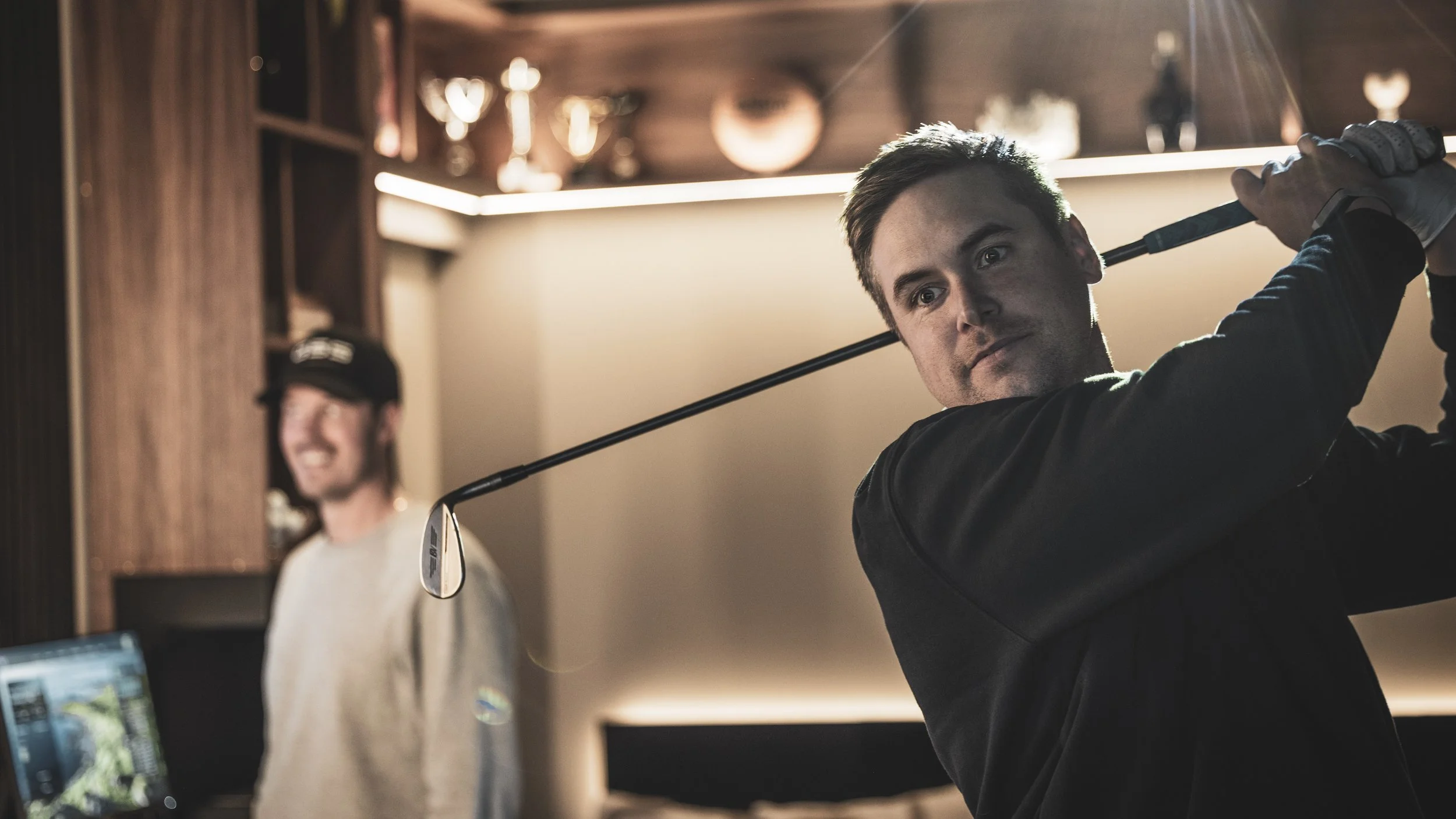 En man som svingar en golfklubgar på ett inomhusgolfbanor, med en annan man i bakgrunden som ler.