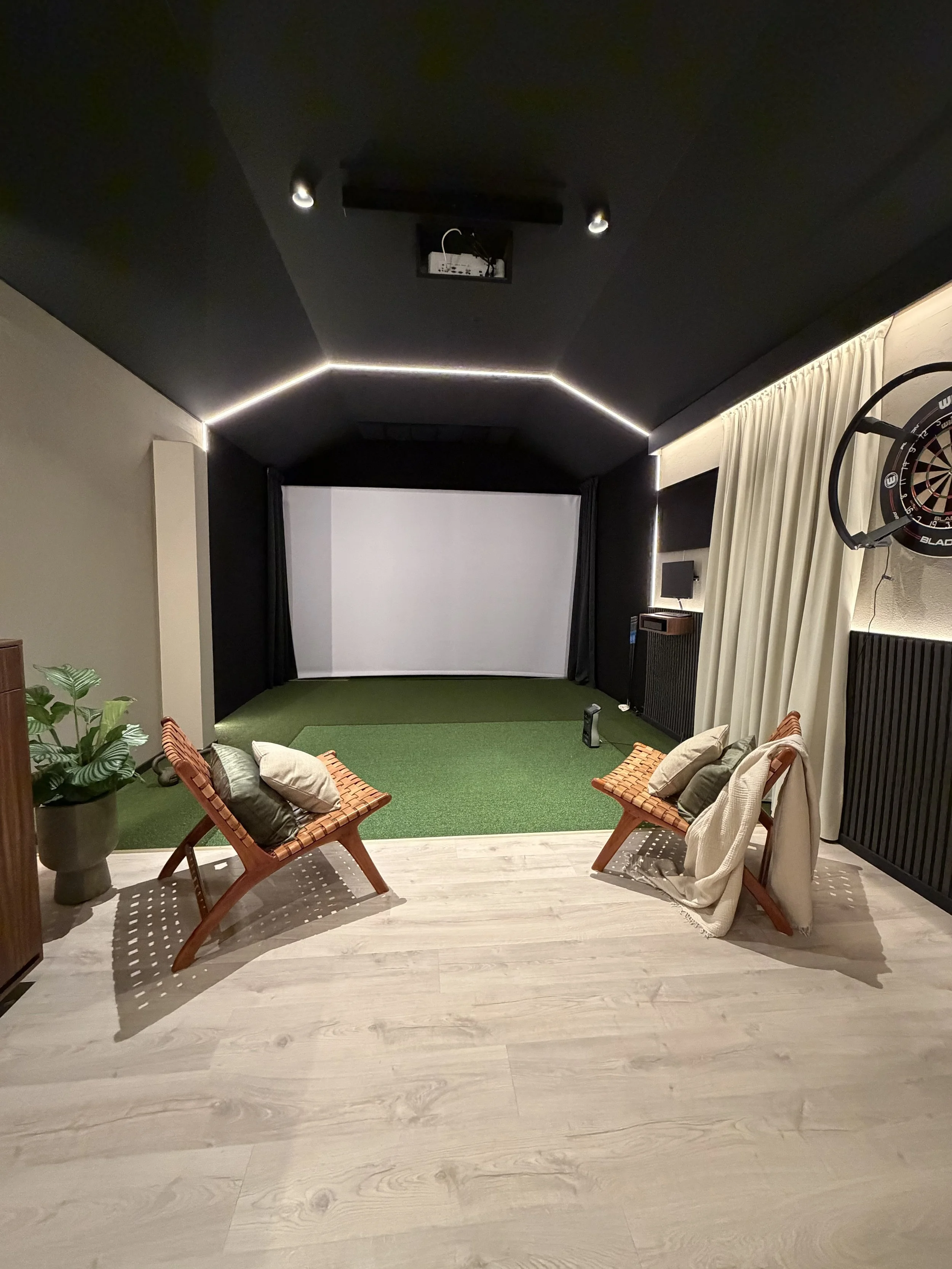 En hemmabio med två stolar framför en stor vit projektorduk, väggarna är svarta och taket är också svart med LED-ljus längs kanterna. Det finns också en darttavla på höger sida och en stor växt på vänster sida.