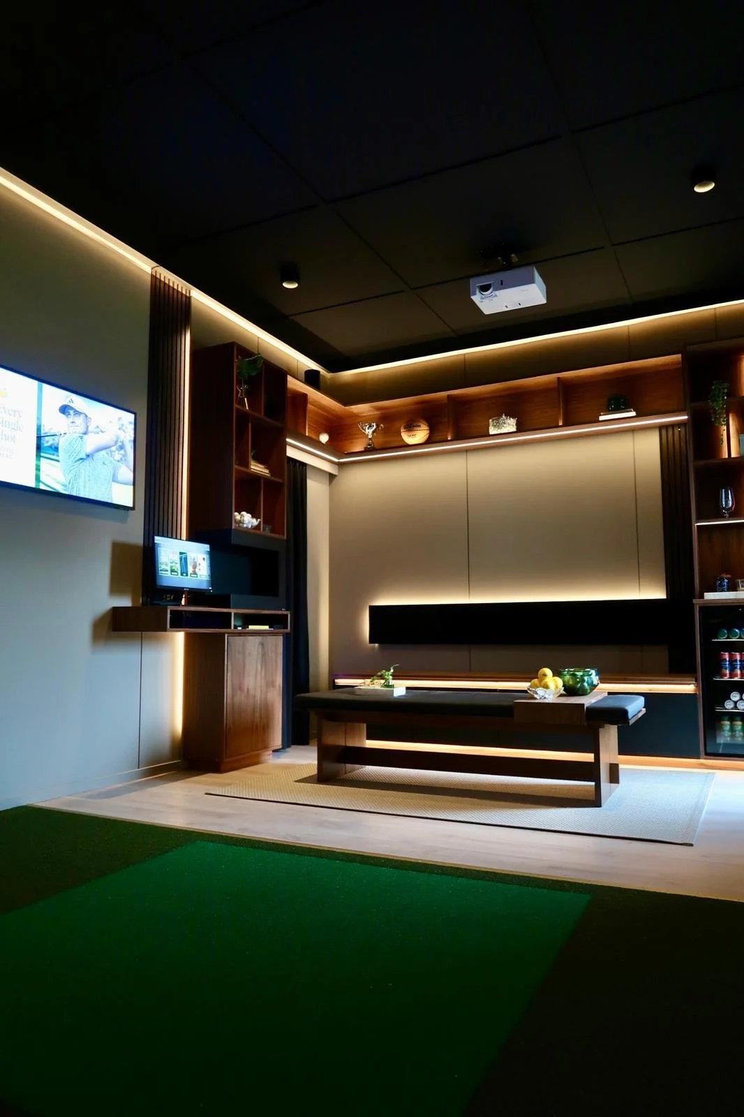 Ett modernt inomhusrum med väggmonterad TV, trähyllor med prydnadsföremål, ett bord med frukt, en vägg med inbyggd belysning och en projektor i taket.