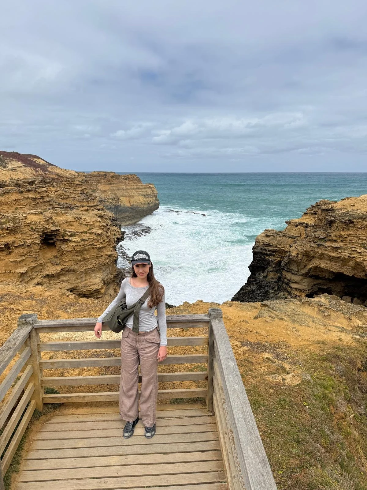 A woman standing on a wooden platform near rocky cliffs overlooking the ocean on a cloudy day.