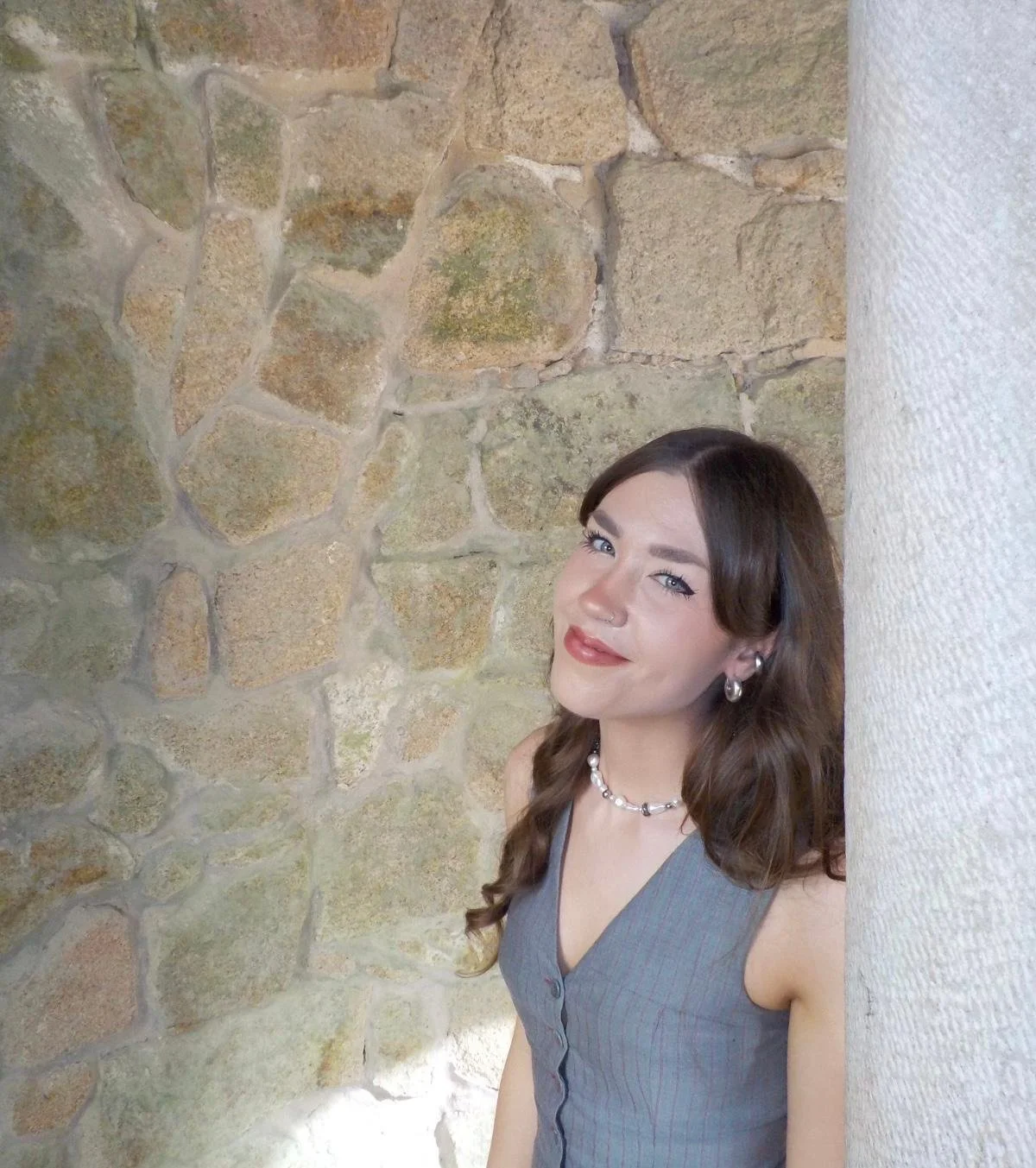 A woman with long brown hair, wearing a gray sleeveless blouse, silver hoop earrings, and a pearl necklace, standing next to a white textured column and a stone wall.