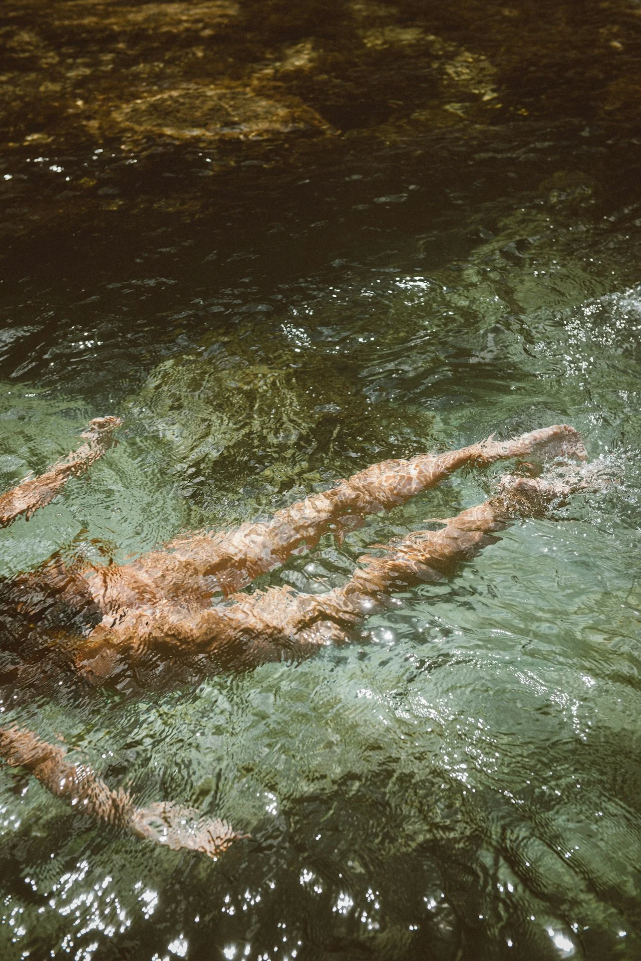 Long, submerged wooden logs floating in a clear, shallow greenish stream with ripples and sunlight reflections.