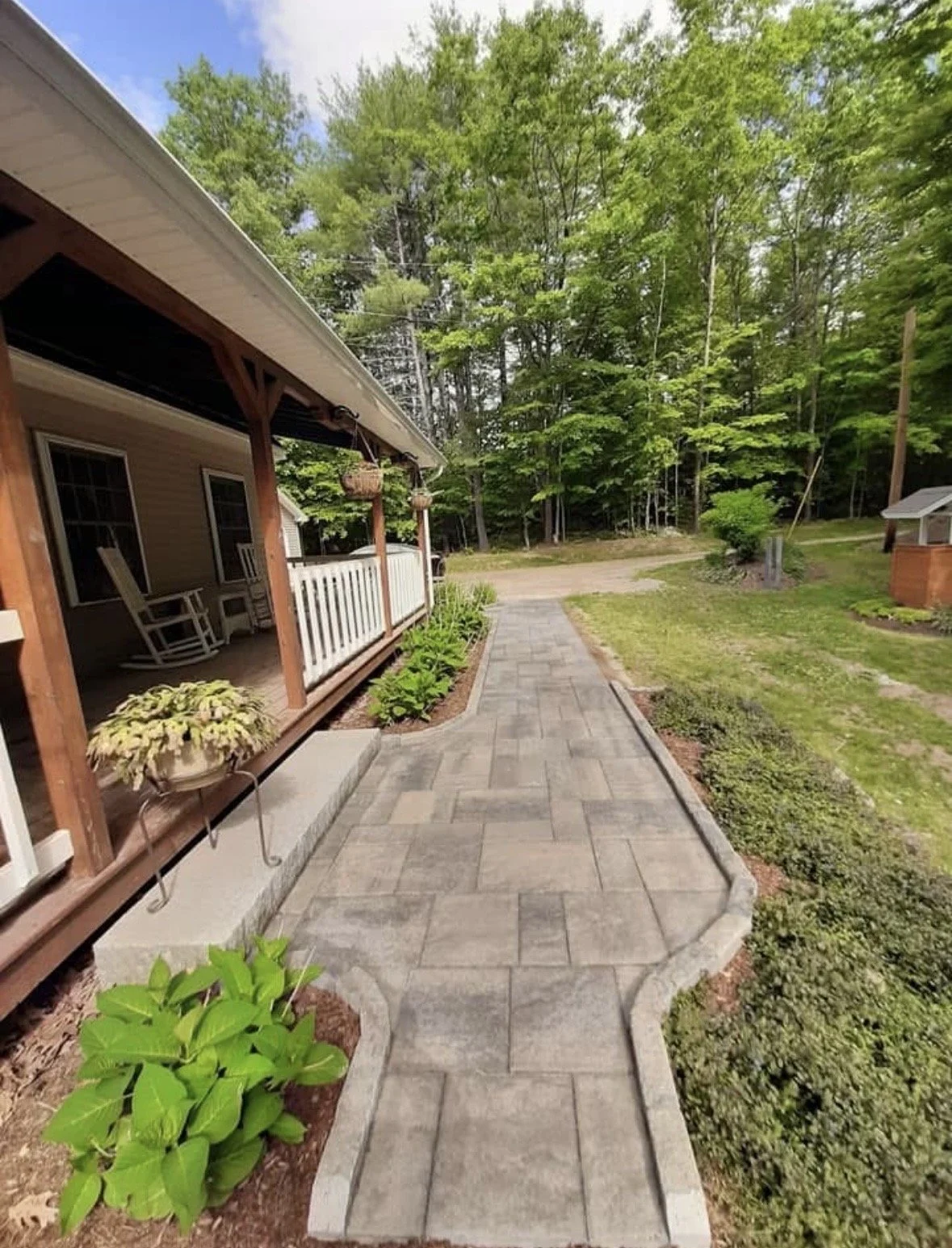 A paved sidewalk leads away from a house on the left, with a front porch featuring rocking chairs and hanging flower baskets. The yard has green plants and hedges, surrounded by tall trees in the background.