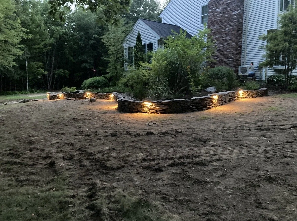 A backyard with a stone retaining wall illuminated by landscape lighting, adjacent to a white house with siding and a brick chimney, surrounded by trees and bushes.