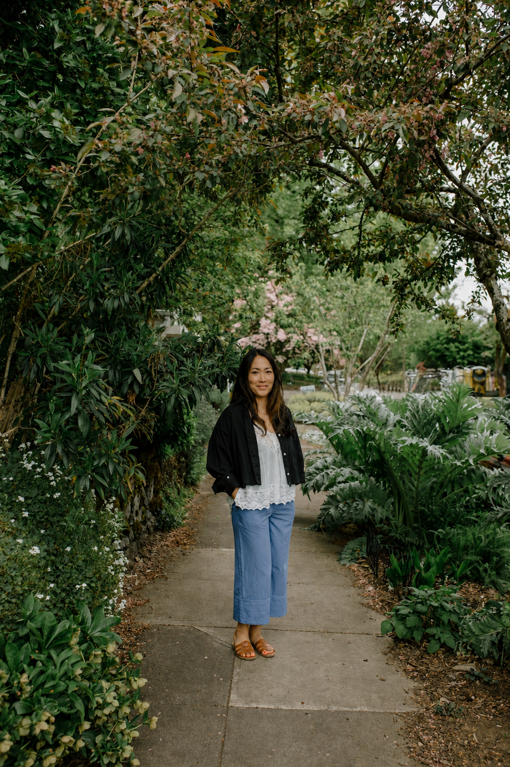 A woman standing on a sidewalk surrounded by lush green plants and trees, smiling at the camera.